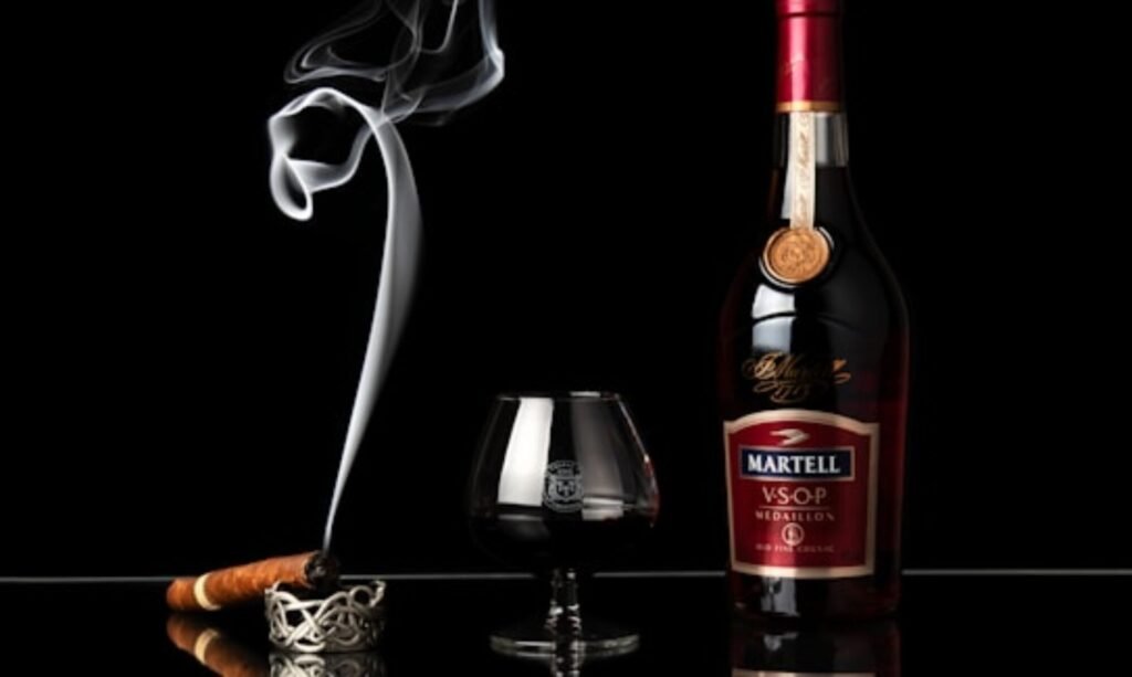 A bottle of Martell VSOP Medaillon cognac stands alongside a glass filled with the dark liquid. To the left of the glass, a lit cigar rests on an ornate ashtray, with a swirl of smoke rising elegantly into the air. The background is completely black, emphasizing the rich colors and reflective surface beneath the objects.